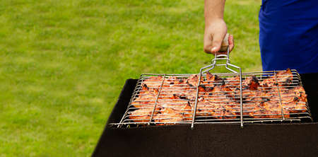 A man is frying kebabs on a green lawn. A mans hand holds a grill with grilled meat in nature. The process of cooking shish kebab.の写真素材