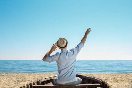 A young guy in a hat relax on the beach, he is joy becasue at lest he is reached the sea. He is sitting on the old boat with hands up.の写真素材