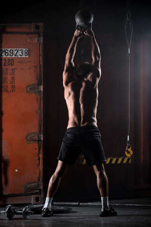 Beautiful athlete doing kettlebell swings. View from the back. Athlete bare-chested. Studio shot in a dark tone.の写真素材