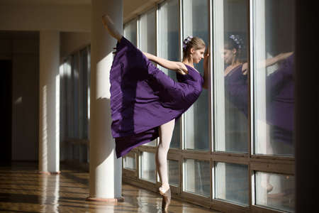 Graceful ballerina dancing in a purple dress leg lifted high, standing on pointe near a large window in the setting sun.の写真素材