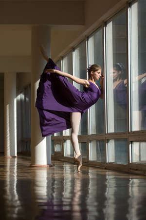 Graceful ballerina dancing in a purple dress leg lifted high, standing on pointe near a large window in the setting sun.の写真素材