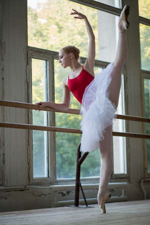 Young ballerina standing on one leg on your toes in pointe and doing stretching, lifting the leg up high. Model holds hands behind the barre.の写真素材