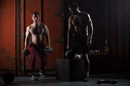 Two beautiful athlete train. One does kettlebell swings, squats with dumbbells other. The picture in the studio in a dark tone.の写真素材