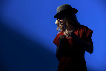 Contrast portrait of sexy blonde in a red fitted dress and a hat that covers her face in shadow.の写真素材