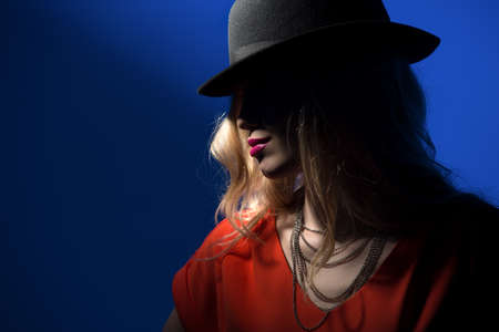 Contrast portrait of sexy blonde in a red fitted dress and a hat that covers her face in shadow.