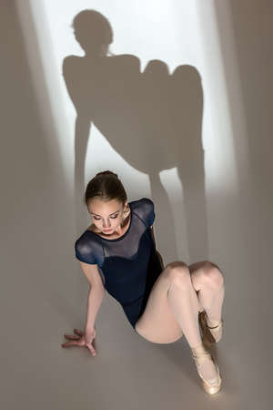 Full-length portrait sitting on the floor of a graceful ballerina in a studio on a white background. Dancer in a blue bathing suit. Against the background of her beautiful shadow.の写真素材