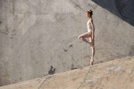 Slim dancer stands in a ballet pose and looking down on a gray urban concrete background. Outdoors shooting with sun light.の写真素材