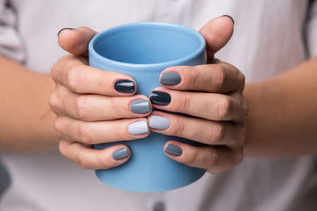 Beautiful well-groomed female hands with a stylish modern manicure holding a blue cup close-up. Each nails are made up of different shades of gray varnish.の写真素材