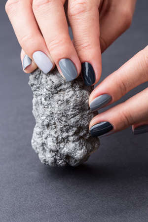 Well-groomed female hands with a stylish manicure holding a beautiful textured silver mineral. Nail painted gray paint different tonality. Grey neutral background.の写真素材