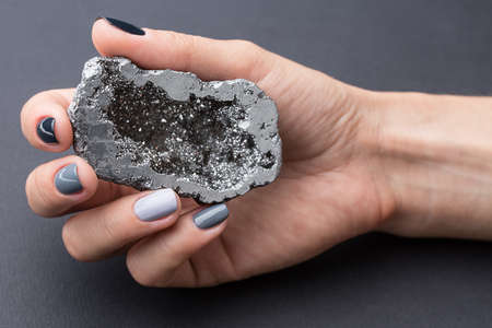 Well-groomed female hand with a stylish manicure holding a beautiful textured silver mineral. Nail painted gray paint different tonality. Grey neutral background.の写真素材