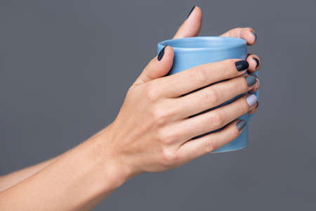 Beautiful well-groomed female hands with a stylish modern manicure holding a blue cup close-up. Each nails are made up of different shades of gray varnish. The isolated image on a neutral gray background.の写真素材