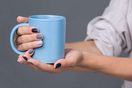 Beautiful well-groomed female hands with a stylish modern manicure holding a blue cup close-up. Each nails are made up of different shades of gray varnish. The isolated image on a neutral gray background.の写真素材