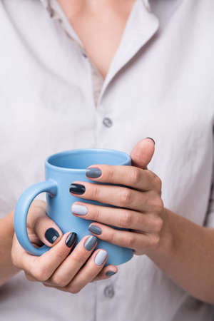 Beautiful well-groomed female hands with a stylish modern manicure holding a blue cup close-up. Each nails are made up of different shades of gray varnish.の写真素材