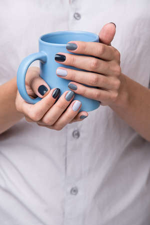 Beautiful well-groomed female hands with a stylish modern manicure holding a blue cup close-up. Each nails are made up of different shades of gray varnish.の写真素材