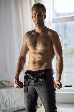 Handsome guy topless with jeans unbuttoned playfully holding a whip BDSM. The picture in the studio...