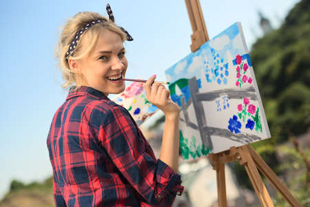Beautiful blonde woman artist in a playful mood dressed in a plaid shirt. She paints a picture with a brush and paints natureの写真素材