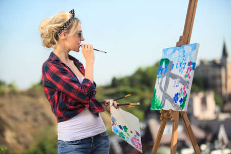 Beautiful blonde woman artist in a playful mood dressed in a plaid shirt with sunglasses. She paints...
