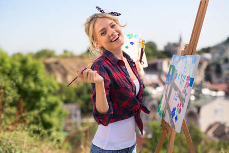 Beautiful blonde woman artist in a playful mood dressed in a plaid shirt. She paints a picture with a brush and paints natureの写真素材