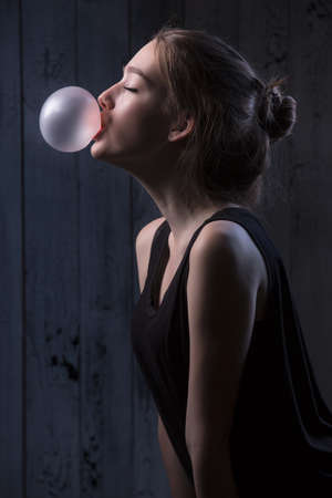 Portrait of sexual brown-haired, which inflates languidly pink bubble of chewing gum. Photographed in studio on a dark background.の写真素材
