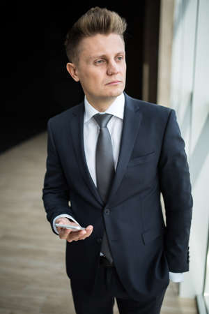 Man in a dark suit standing with phone in hand. Thinking and looking out the window.の写真素材