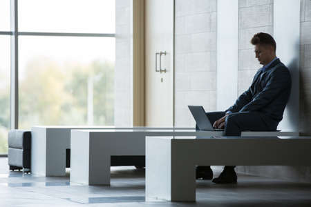 Blonde business man in a dark suit sitting on a bench at a laptop. Horizontal photoの写真素材