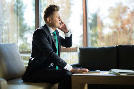 Business man in a dark suit talking on the phone. In the lobby of the hotel sitting on the couchの写真素材