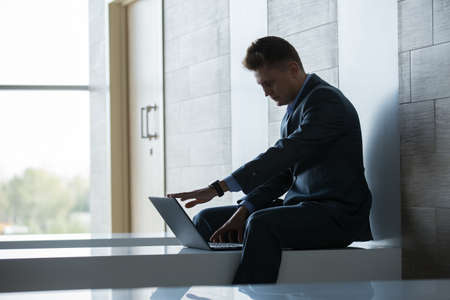 Business man in a suit sitting on a bench against the wall. It works at a laptop. Horizontal close-up photoの写真素材