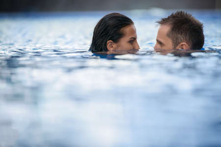 Unusual tanned young couple indulges in a swimming pool. The dark-haired girl and boy looking at each other's eyes downcast face half submerged. Tropics.の写真素材