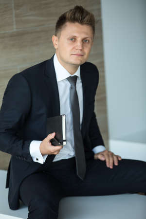 Brown man in a dark suit and an expensive black tie. He sits on a white bench. Keep the black notebook in his hand.の写真素材
