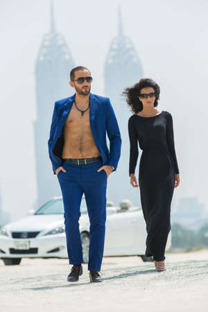 Tall couple came out of the white car. A walk on a deserted beach in Dubai on the background of skyscrapers. The dark-haired woman in high heels with a fluffy head of hair in a black dress. A tall man in a blue suit with a naked torso. UAE. Vertical photoの写真素材