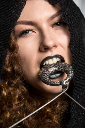 Interesting art studio shot curly girl in a black hood and dark lips. Teeth holds a round metal...