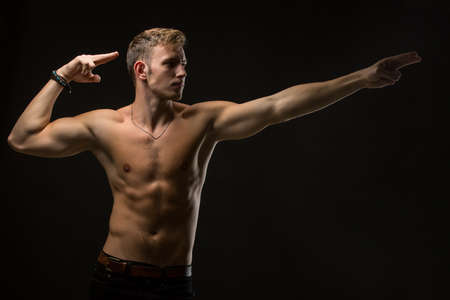 Enviable man with a naked torso and black pants. In the studio on a black background. In a twist. It shows in side arms. Horizontal photoの写真素材
