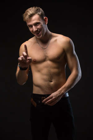 Smiling man with a naked torso and black pants. In the studio on a black background. Buttoning his belt. Finger waves - no. Vertical photoの写真素材