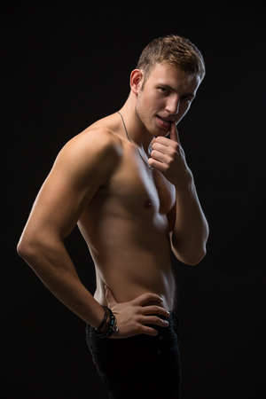 Sexy young man with perfect athletic body. The black pants with a naked torso. Cute a finger on his lips. Vertical photoの写真素材