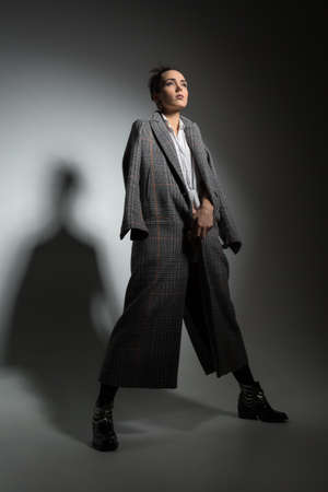 Girl in full growth standing on a gray background in a studio. She is standing with her legs apart and holding her hands together in front of her. She dressed in a long coat with patterns. Shoot in a low key. Vertical.の写真素材