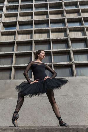 Pretty ballerina posing on the concrete wall background. She holds her hands on the belt while one leg stands on the toe. She dressed in the black dance suit with black tutu and pointe shoes. Full growth photo outdoors. Vertical.の写真素材
