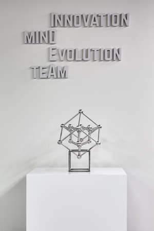 White cubic stand with metallic atom model on the white wall background. There is a volumetric gray...