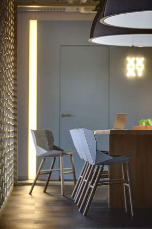 Table with a laptop on it and chairs standing near the table. Two large lamps over the table. On the...