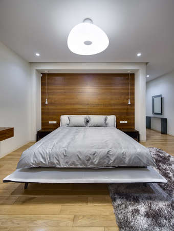 Bedroom in a modern style with light walls. There is a big bed with gray bedcover and pillows. On...