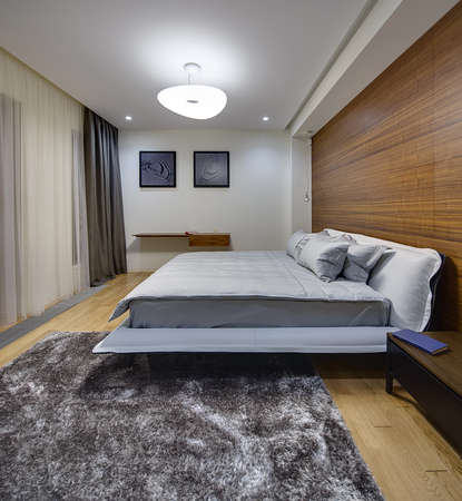 Bedroom in a modern style with light walls. There is a big bed with gray bedcover and pillows. On the right side of the bed there is a dark wooden table with a book. On the right wall there is a big niche with wooden panel and hanging lamp. On the back waの写真素材
