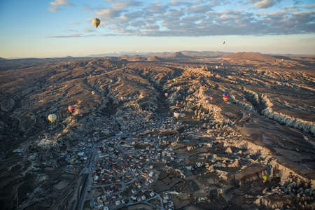Multi-colored air balloons are flying above the town in the valley. Balloons colored in red, white, blue, yellow, orange, green. On the background there is a blue sky with clouds. View from the top.の写真素材