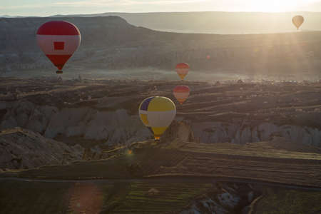 Multi-colored air balloons are flying above a road in the valley. Balloons colored in red, blue, white, yellow, orange, gray. Sun shines from the back. On the background there is a town. View from the side.の写真素材