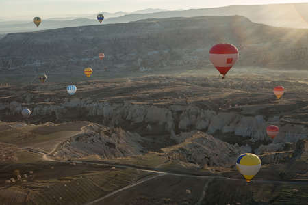 Multi-colored air balloons are flying above roads in the valley. Balloons colored in red, blue, white, yellow, orange, gray. Sun shines from the back.  View from the side.の写真素材