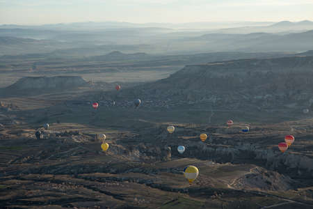 Multi-colored air balloons are flying above roads in the valley. Balloons colored in red, blue, white, yellow, orange, gray. On the background there is a town, sky in a haze and mountains. View from the top.の写真素材