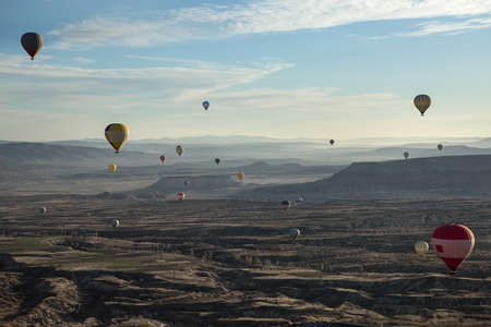 Multi-colored air balloons are flying above the valley on the background of the blue sky with clouds. Balloons colored in red, white, blue, yellow, orange, gray. On the back there is a town and mountains. View from the side.の写真素材