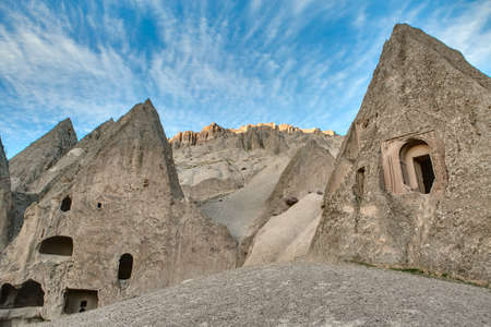 Rock formation with houses carved into rock on the background of the mountains and the blue sky with clouds. Sunlight touches the peaks of mountains at the back.の写真素材