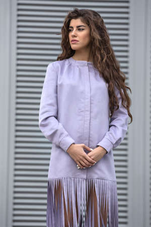 Pretty young girl with amazing hairstyle stands on the striped wall background. She looks to the left while holding her hands together. She wears the design lilac jacket. Vertical.の写真素材