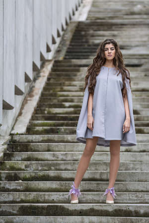 Attractive girl with the beautiful hair stands on the concrete stair. She wears the gray coat and the light shoes with the violet decor. She looks into the camera and holds her hands along the body. Outdoors. Vertical.の写真素材