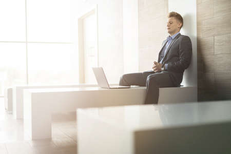 Business man sitting alone on a bench with a laptop inside a building with a modern interior and large windows.の写真素材