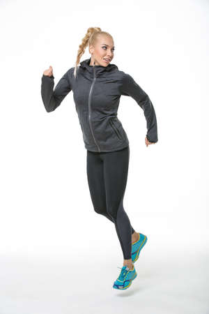 Young blonde girl in the sportswear runs on the white background in the studio. She wears...